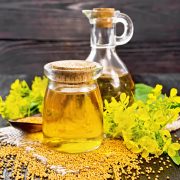 Mustard oil in a glass jar and decanter, mustard grains on a burlap napkin, flowers and leaves on wooden board background